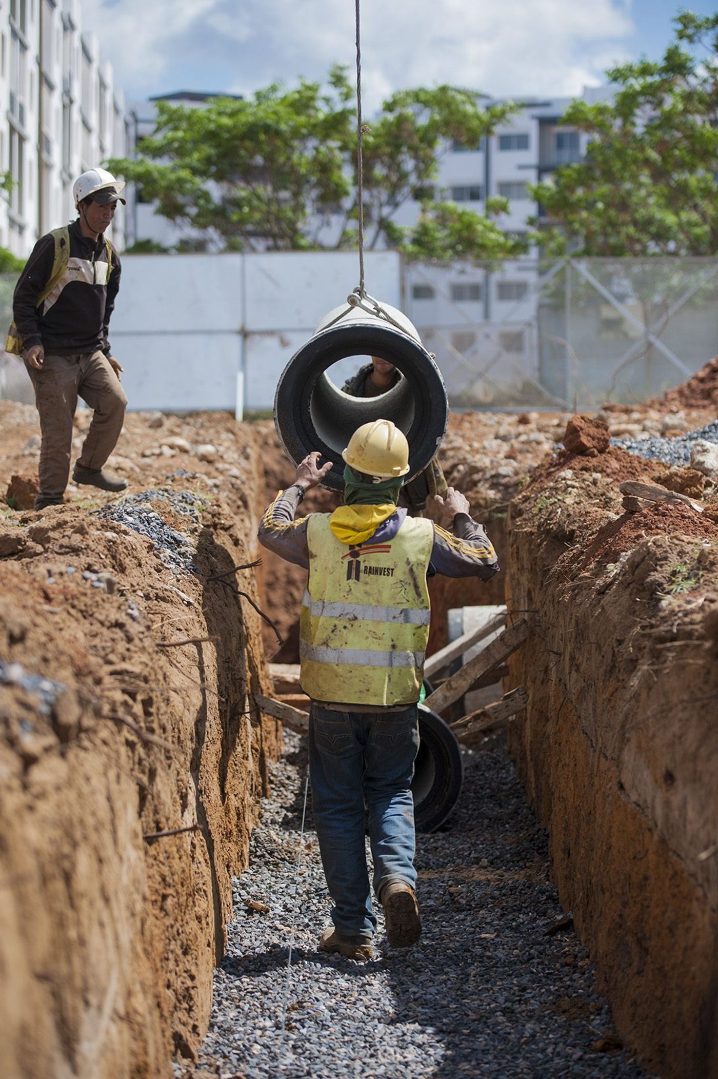 Workers in hard hats lower a large concrete drainage pipe into a dug trench on a construction site