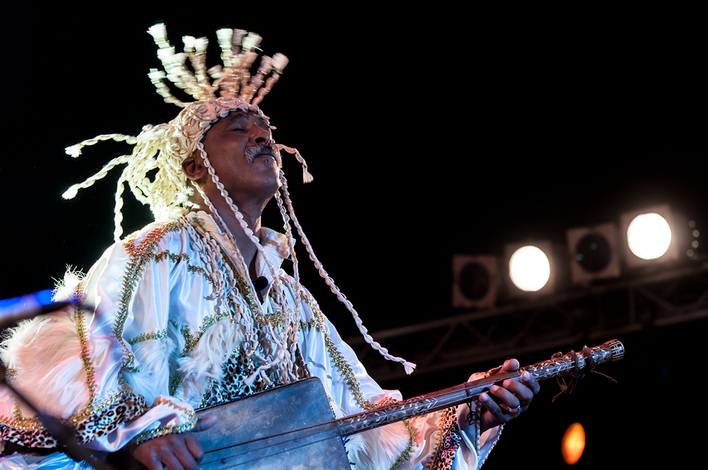 Gnaoua musician in beaded cap and white tunic plays guembri under stage spotlights