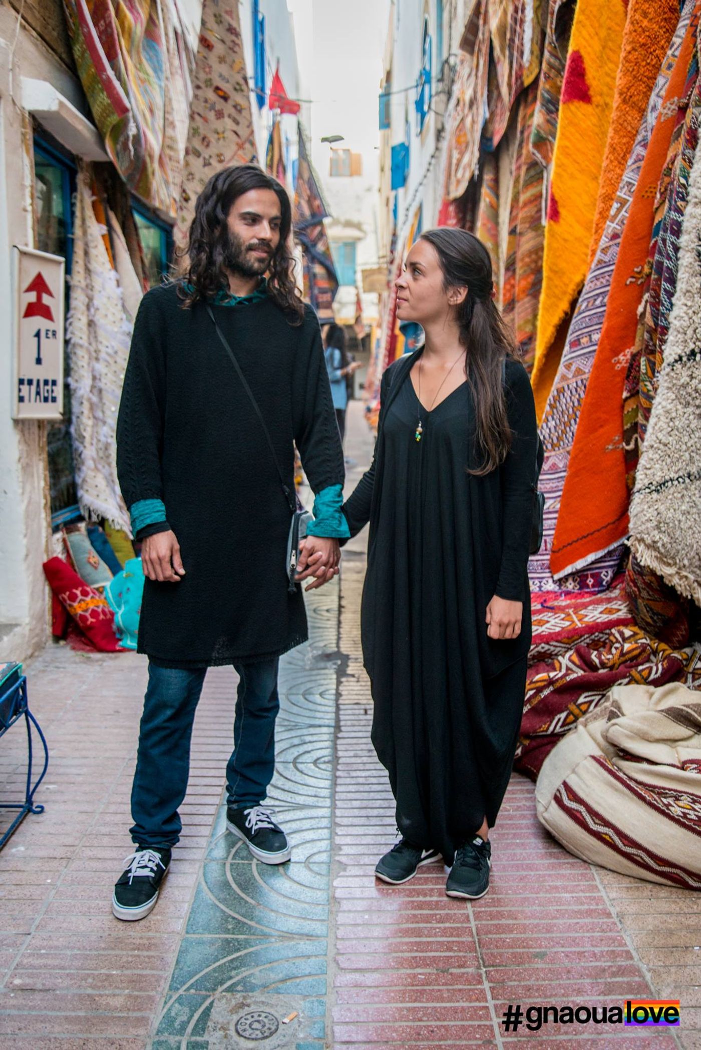 Couple in black holding hands in a narrow Essaouira alley hung with colorful carpets