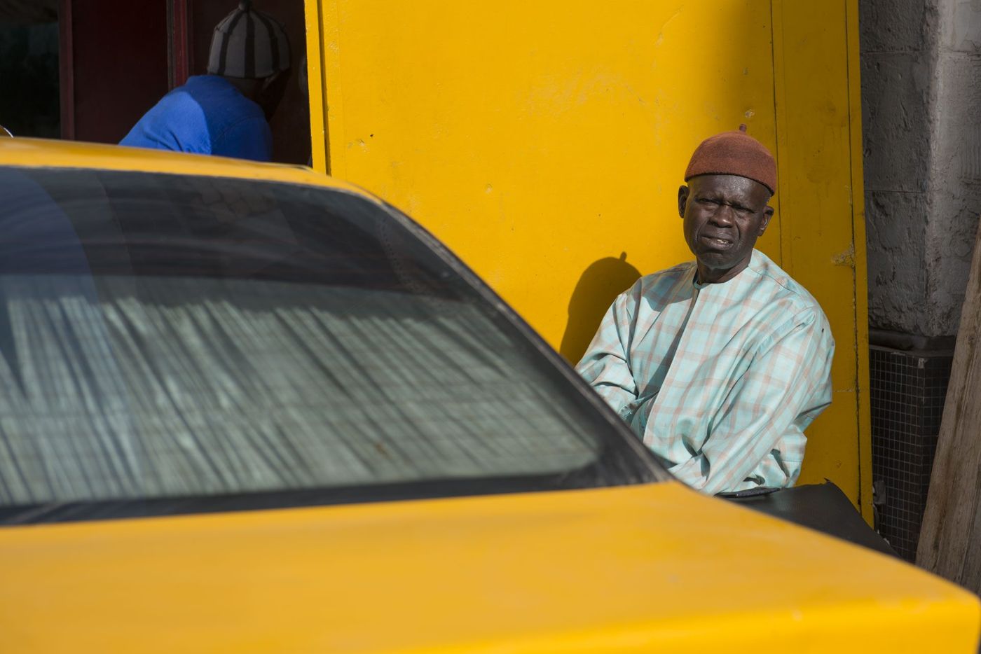Elderly man in pale plaid robe and red fez sits against a yellow wall beside a yellow taxi