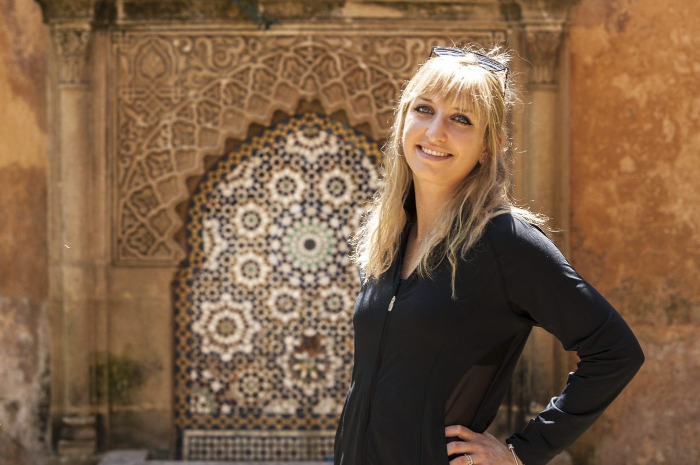 Blonde woman in black jacket smiles before an ornate Moroccan zellige tile fountain