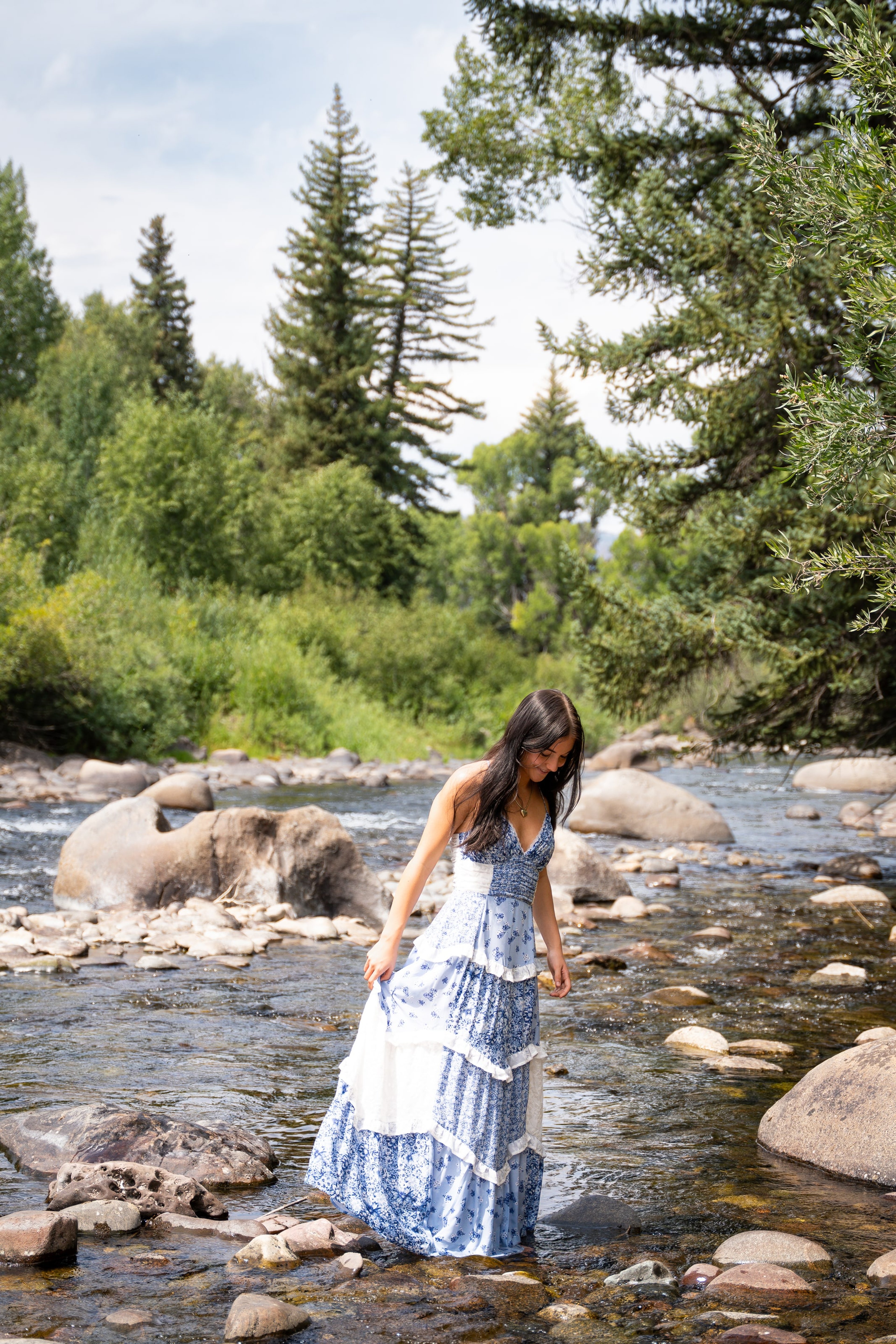 Lucy standing in the creek shallows in a blue dress, pines behind