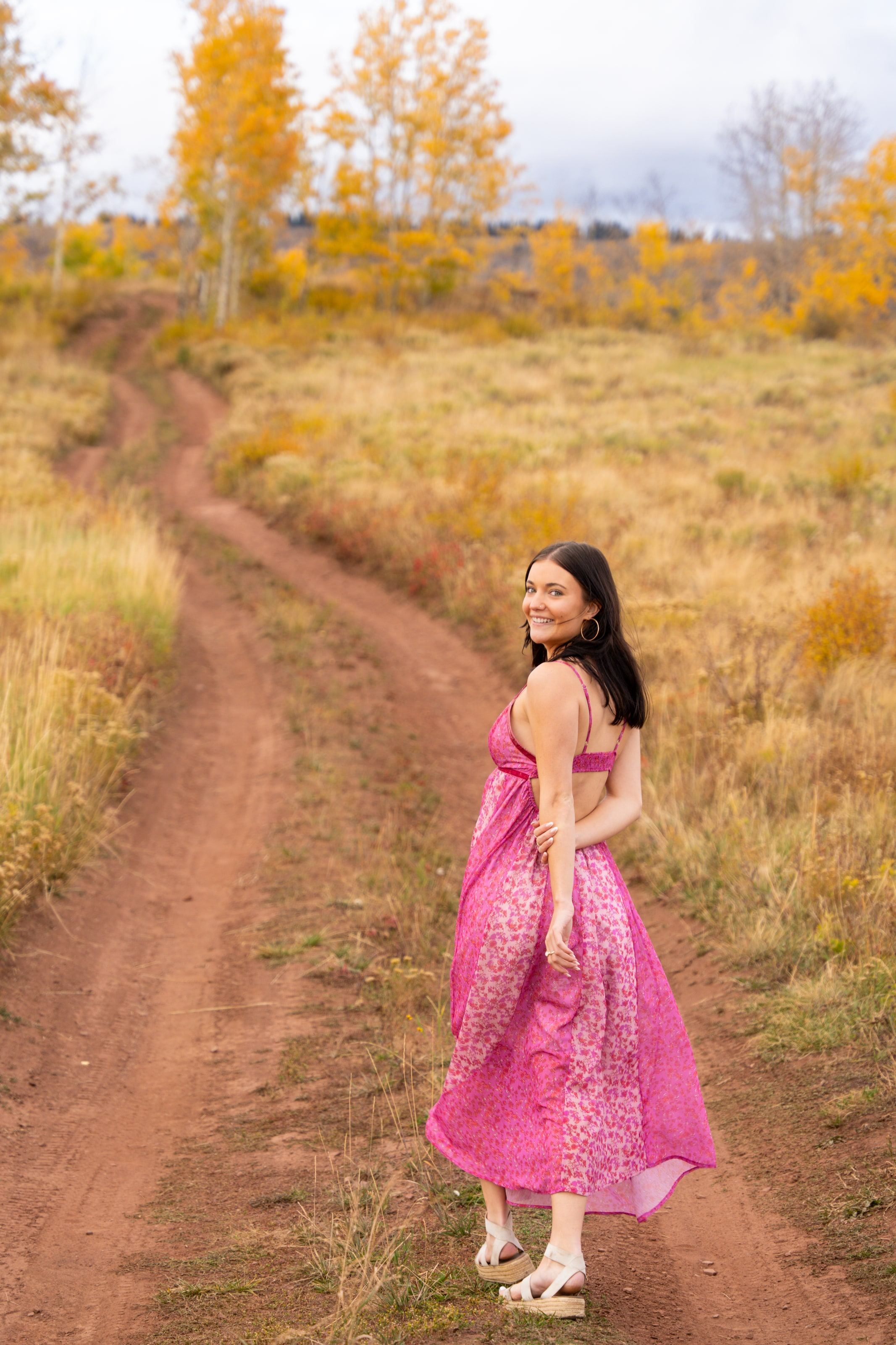 Lucy in a pink dress glancing back on a red dirt road through golden aspens
