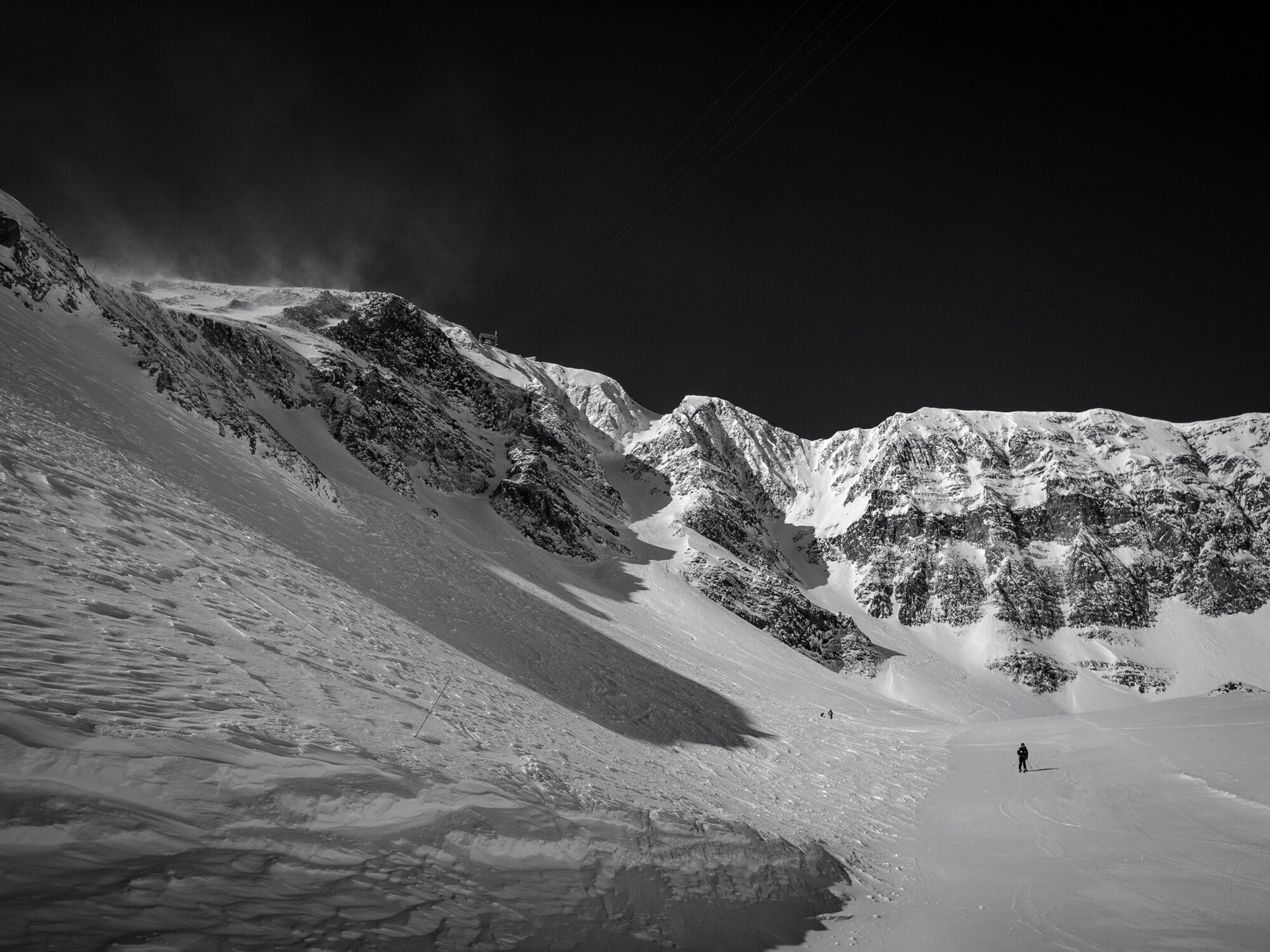 A lone skier crossing the base of a vast snow bowl beneath dark ridgelines, in black and white