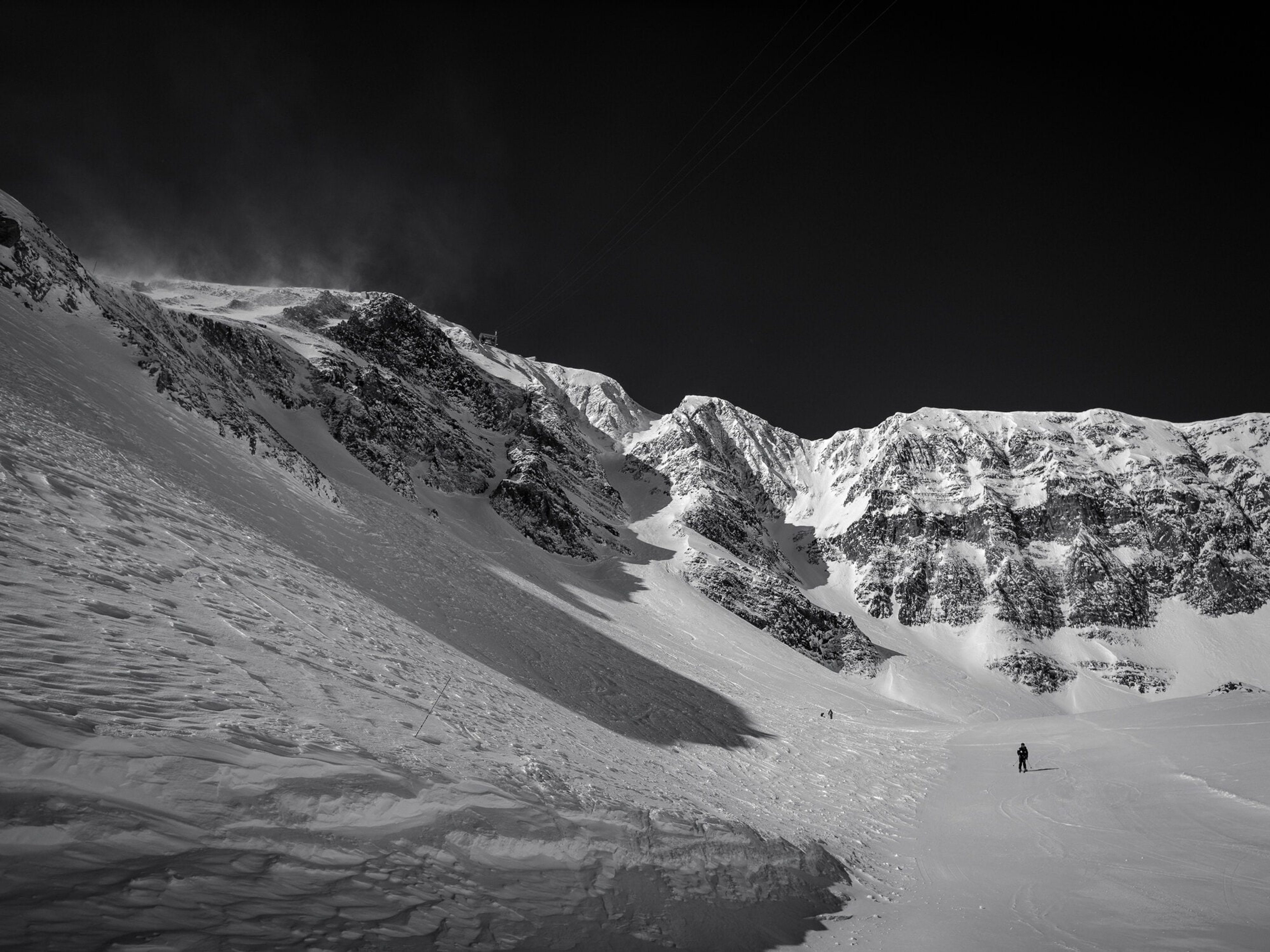A lone skier crossing the base of a vast snow bowl beneath dark ridgelines, in black and white