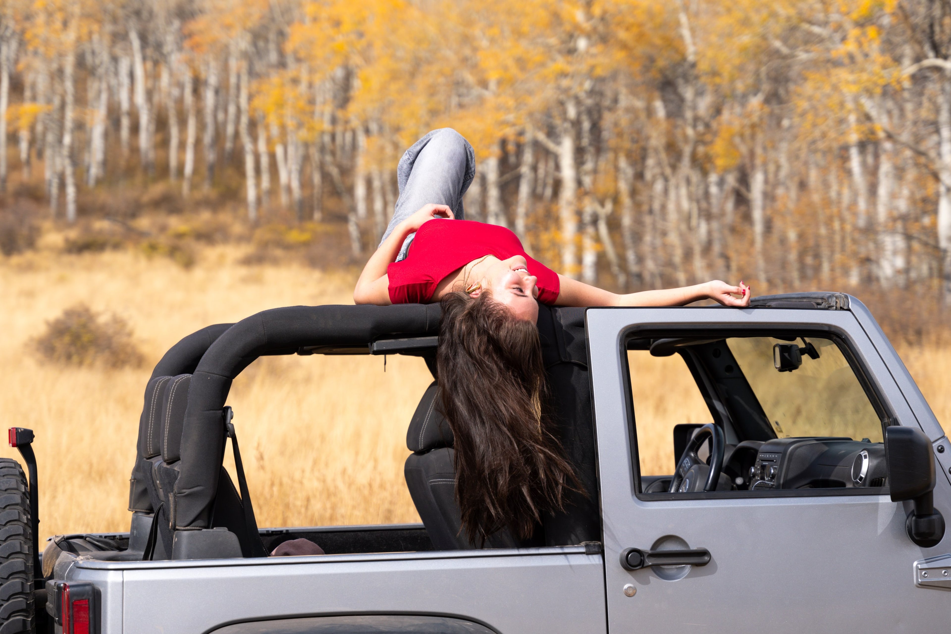 Lucy leaning out of the open Jeep, arms spread, aspens gold behind