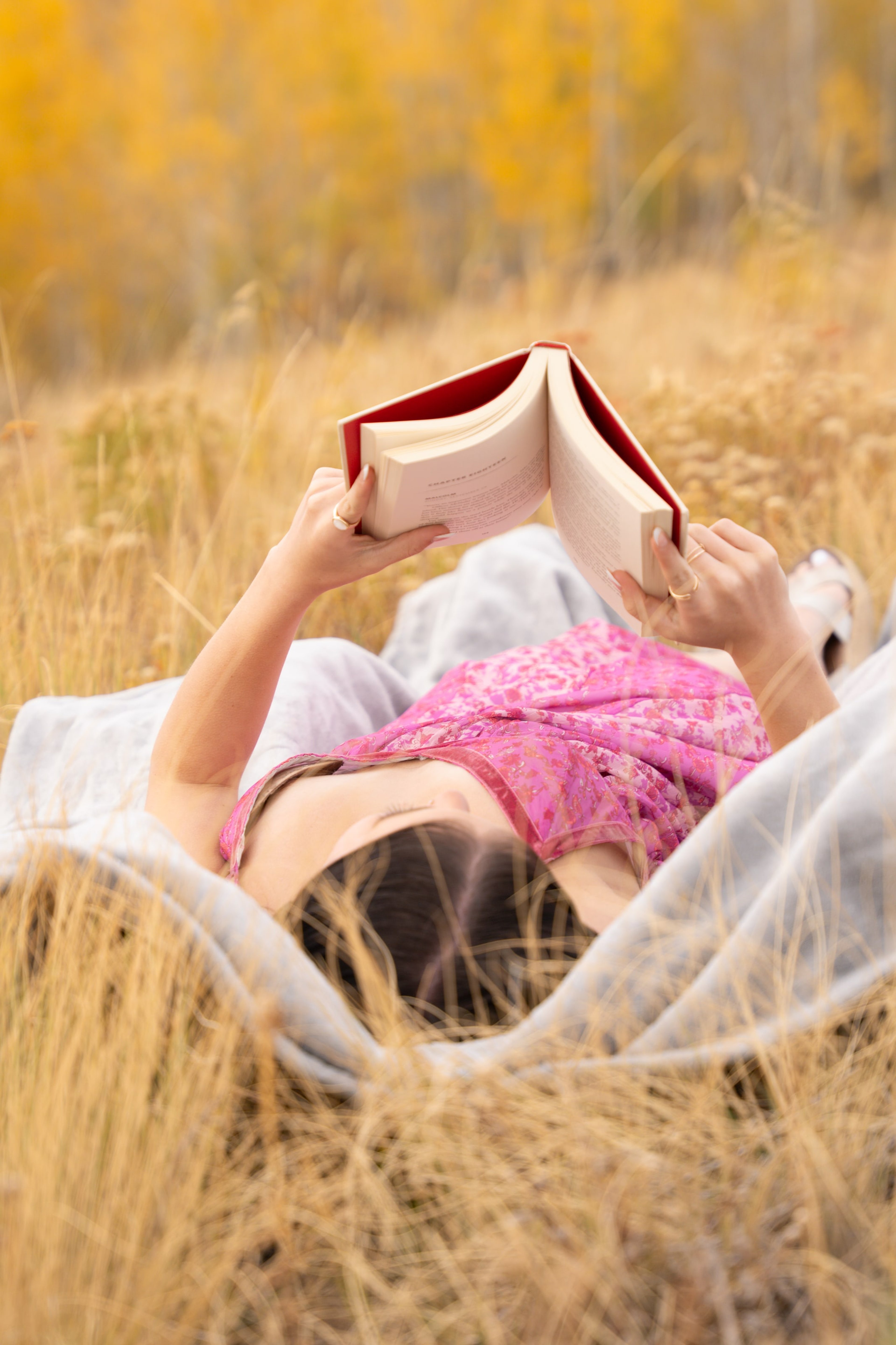 Lucy lying in golden grass reading a red book overhead