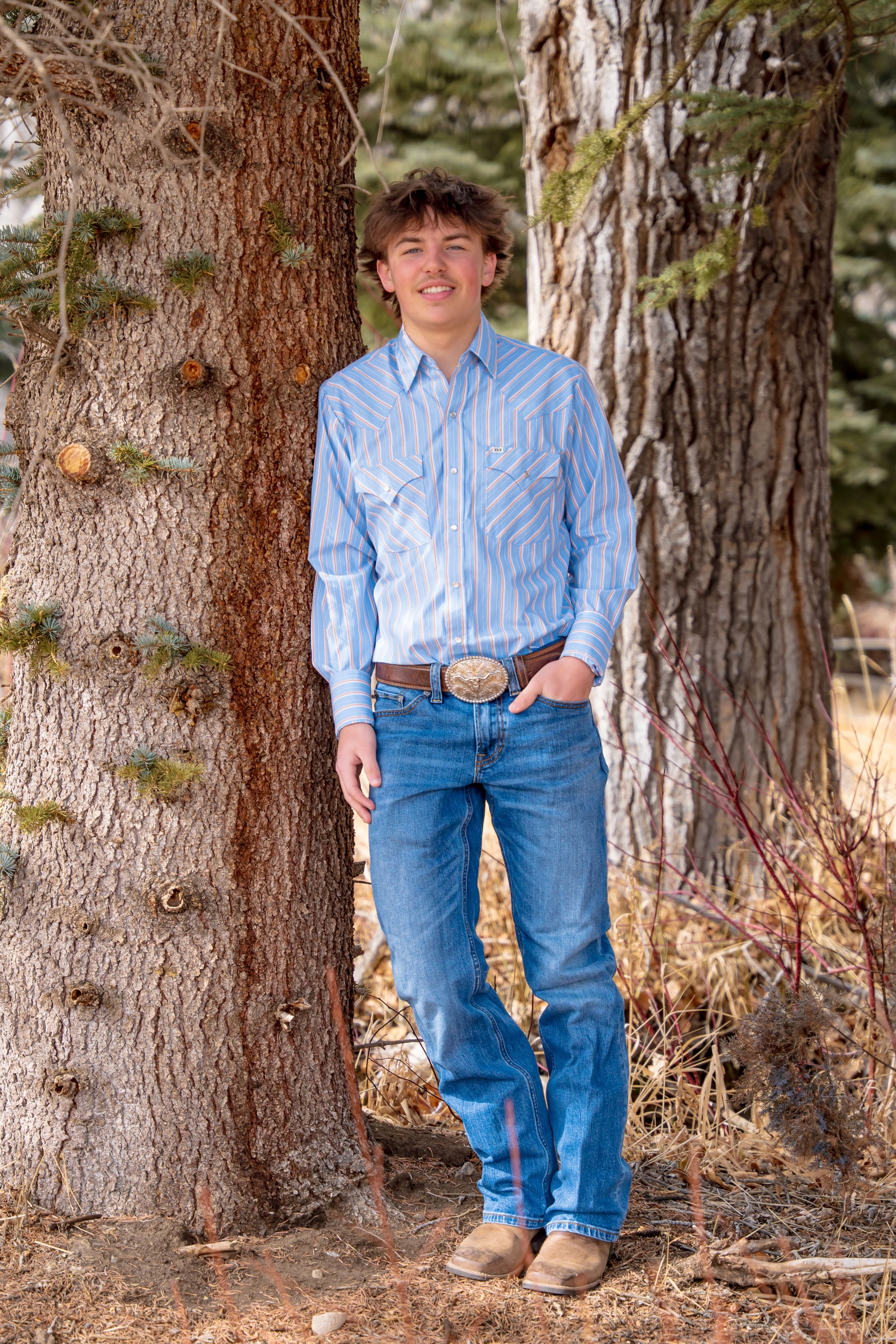 Teagan leaning against a big pine in blue shirt and jeans
