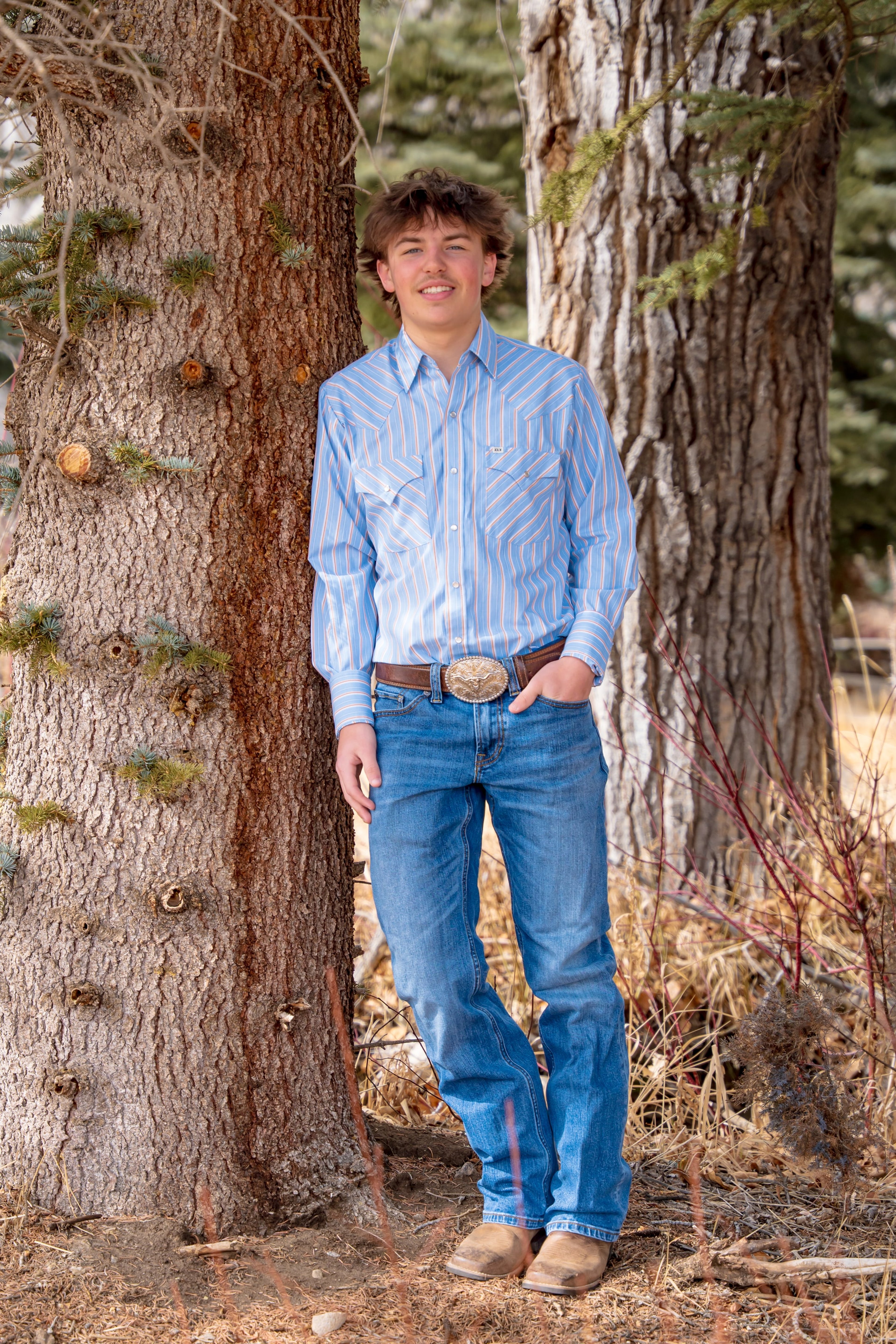 Teagan leaning against a big pine in blue shirt and jeans