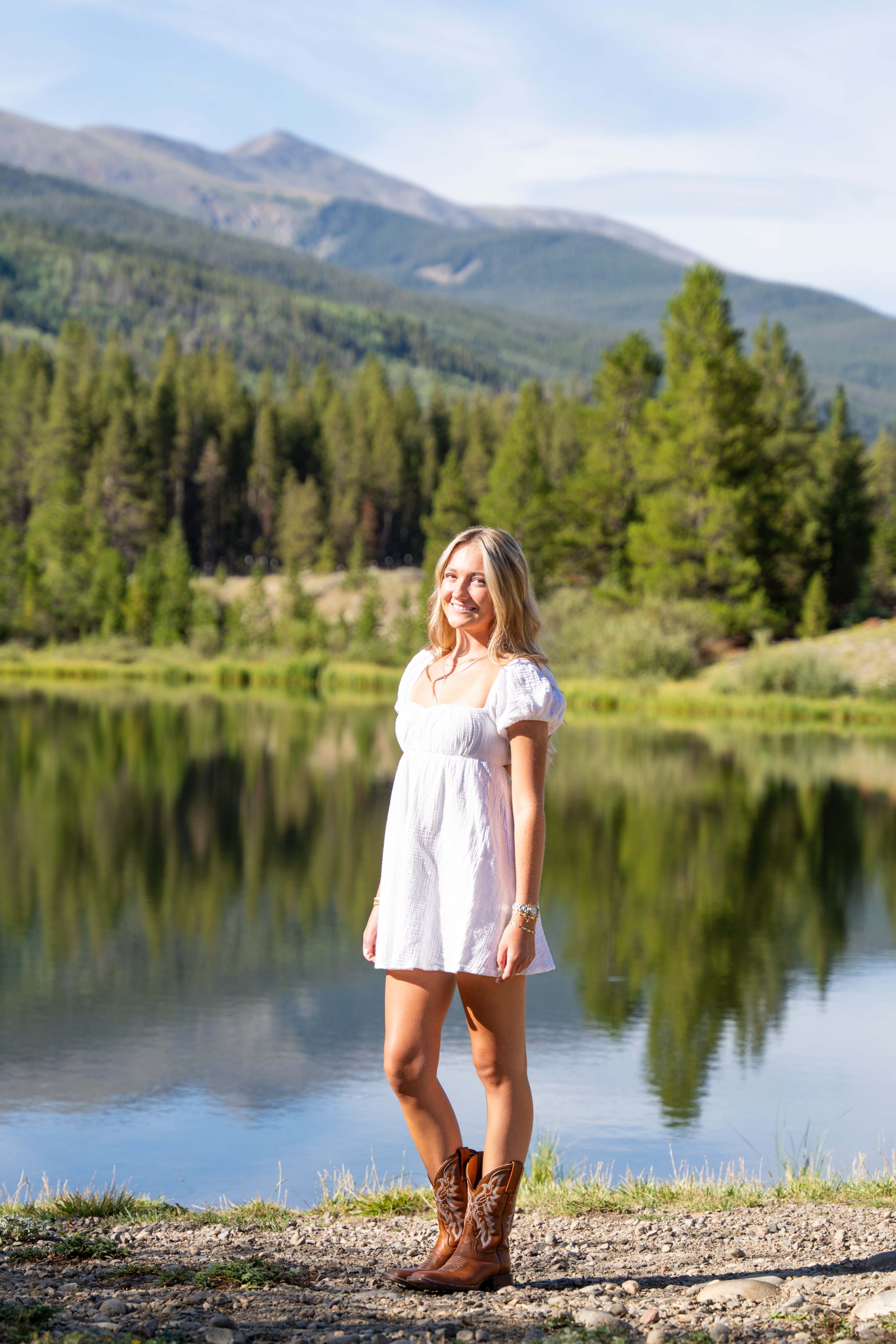 Caleigh in a white dress and cowboy boots beside a lake reflecting forested mountains