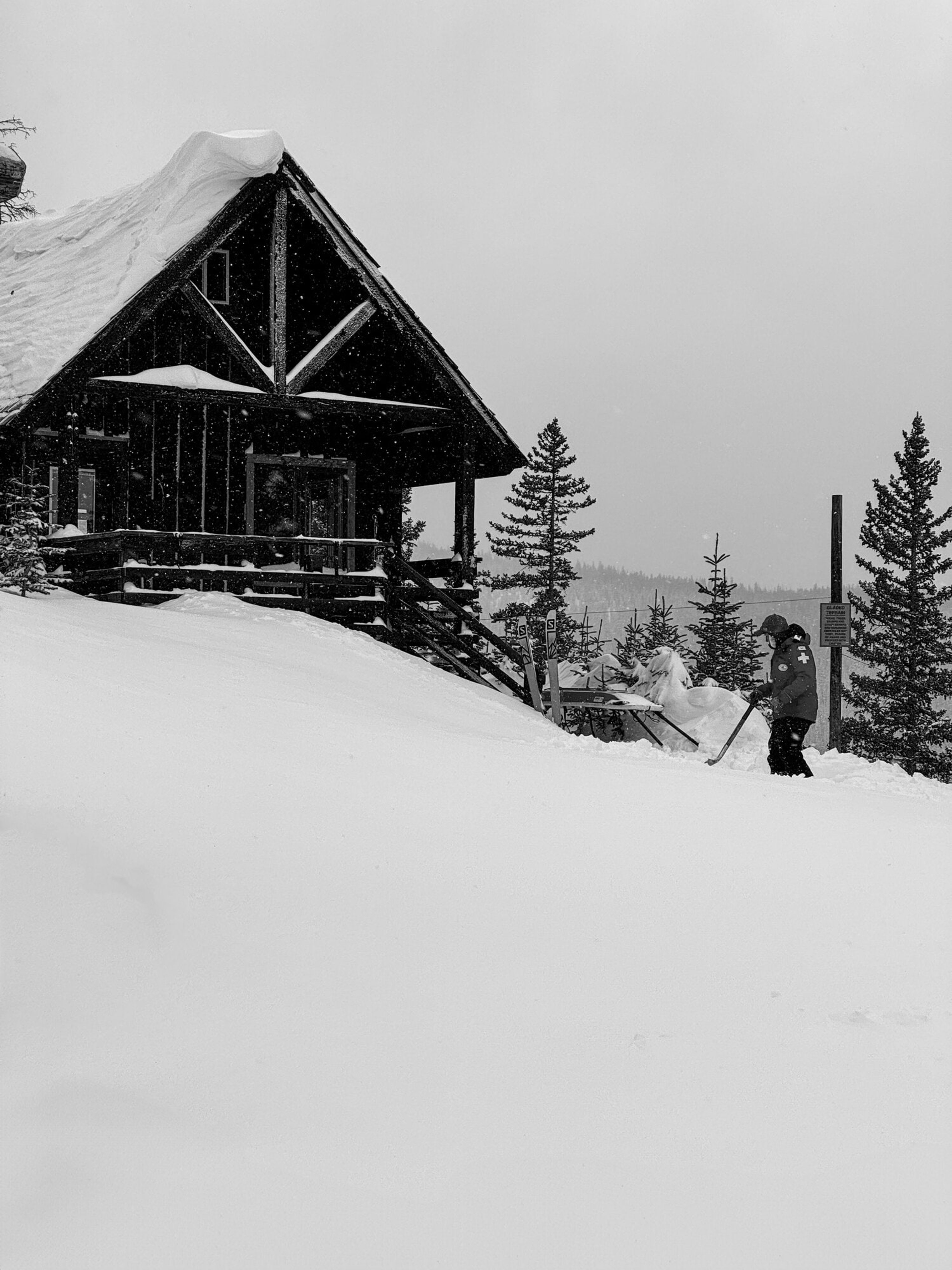 A ski patrol hut buried to its eaves in fresh snow, a skier passing behind, in black and white