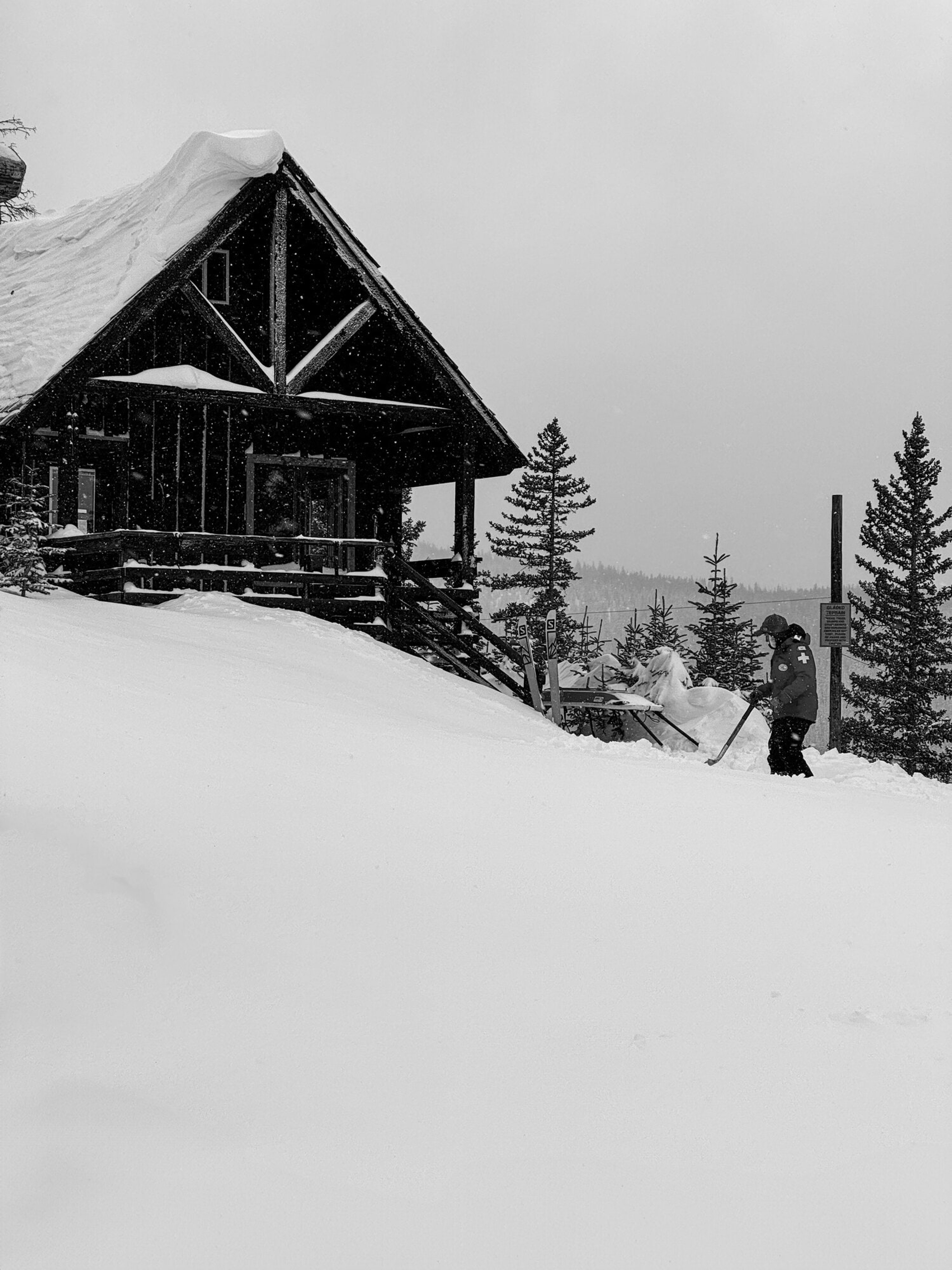 A ski patrol hut buried to its eaves in fresh snow, a skier passing behind, in black and white