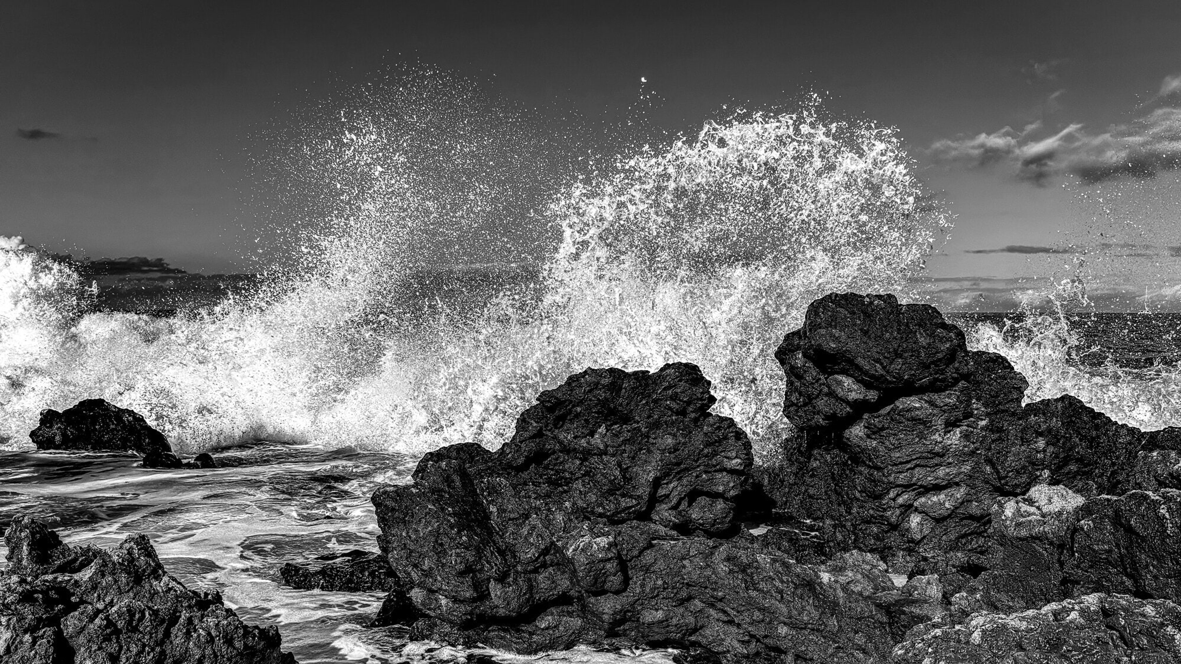 A wave exploding against black lava rock, spray frozen mid-air, in black and white