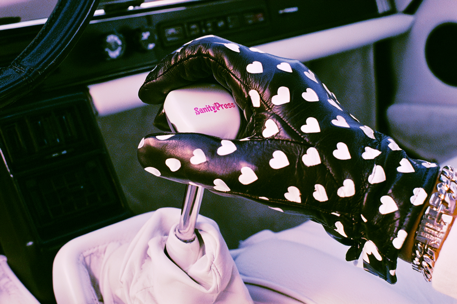 a black leather hand glove with white heart patterns holding a white shifter with the text "SanityPress", inside a vintage porsche white interior, first person perspective, aged technicolor film aesthetic