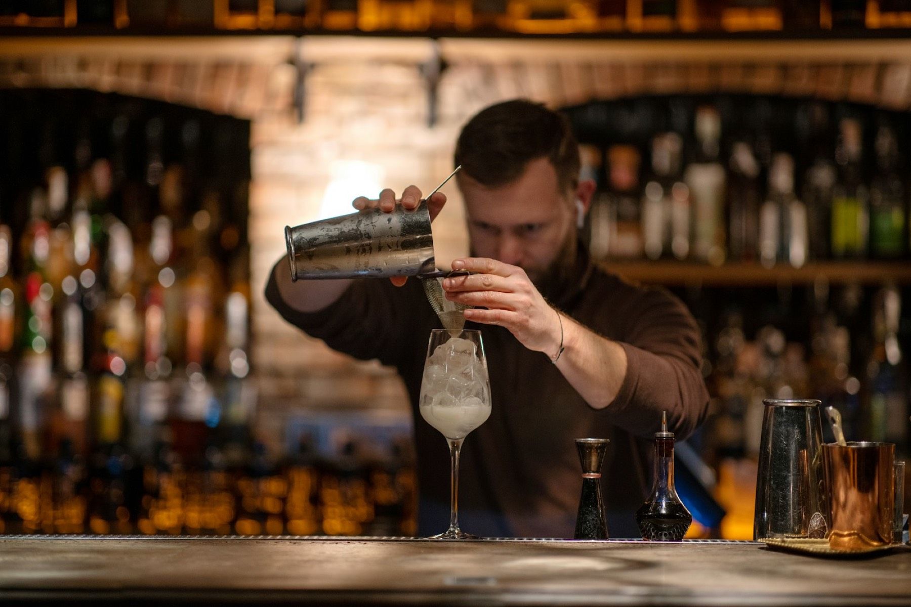 A bartender pours from a shaker into a coupe, a warm back bar of bottles behind.