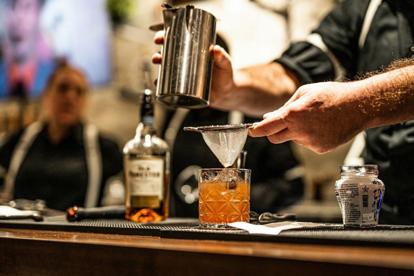 A bartender fine-strains a drink at service, a bottle waiting on the speed rail.