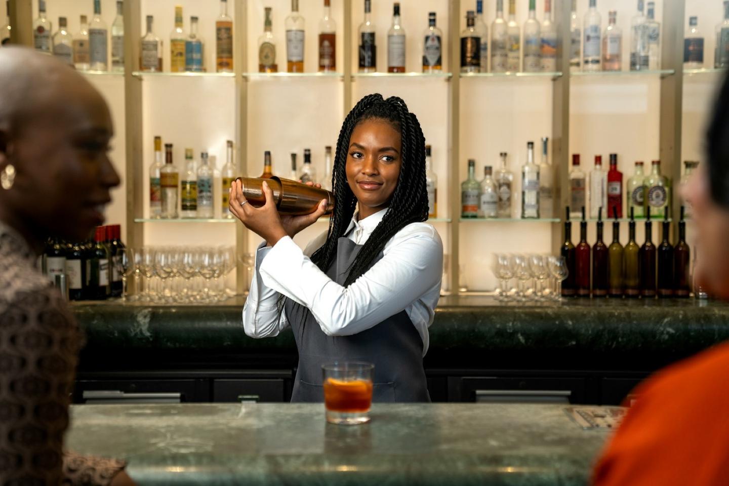 A bartender shakes a drink at a hotel bar, guests at the counter and a lit wall of bottles behind.