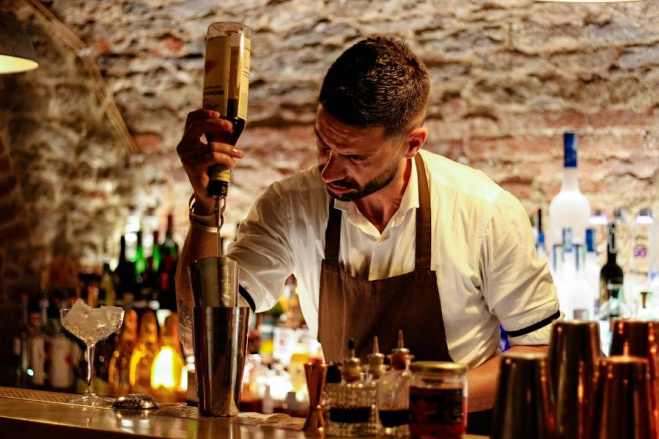 A bartender in an apron pours from a bottle into a shaker tin against a brick wall.