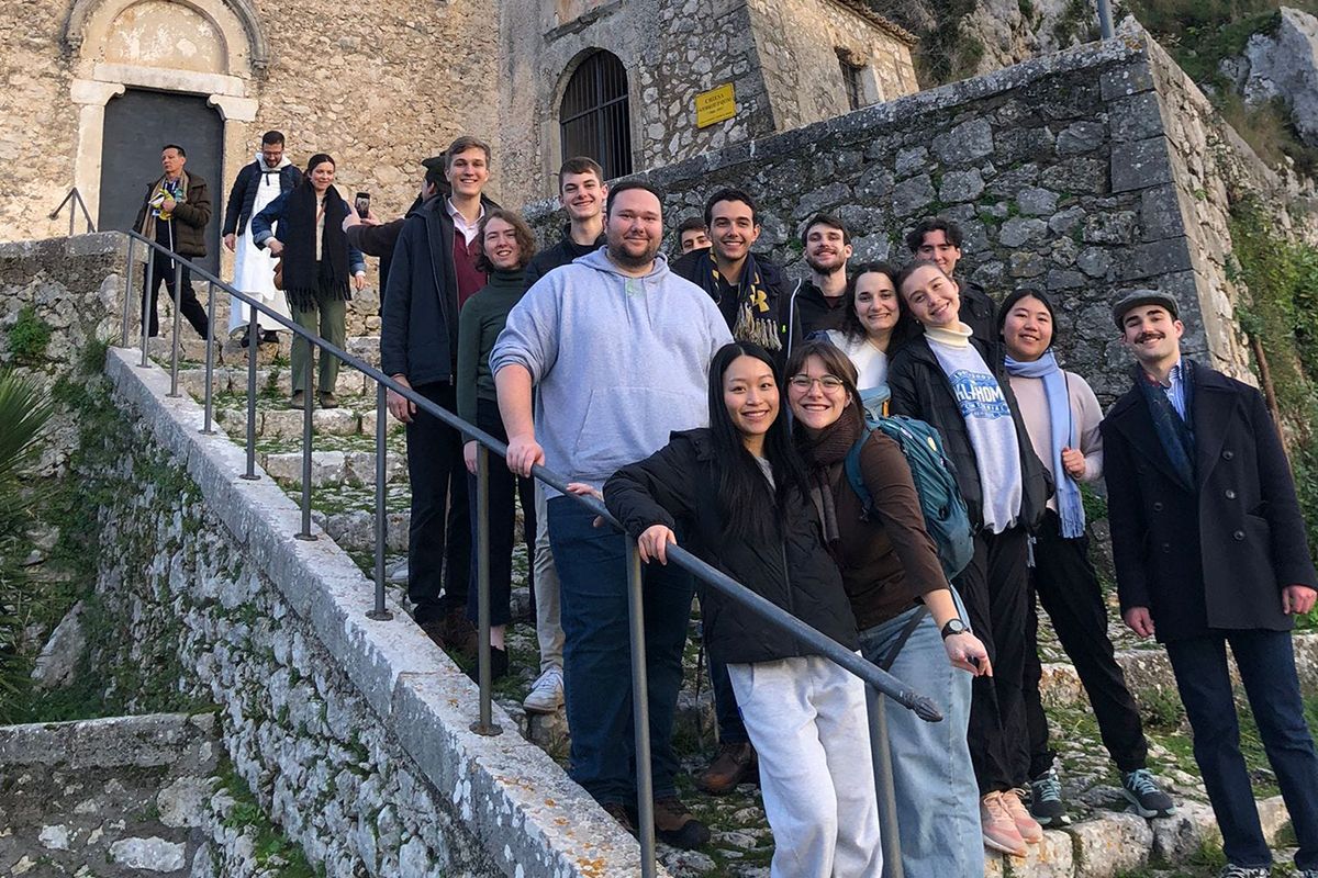 A group of students smile on a stone staircase in front of a stone building.