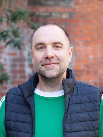 Colin smiling in a head-and-shoulders portrait in front of an old brick wall, framed by foliage.