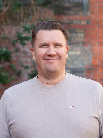 Jarno smiling in a head-and-shoulders portrait in front of an old brick wall, framed by foliage.