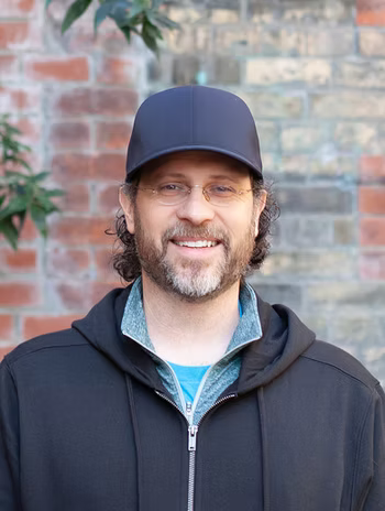Casey smiling in a head-and-shoulders portrait in front of an old brick wall, framed by foliage.