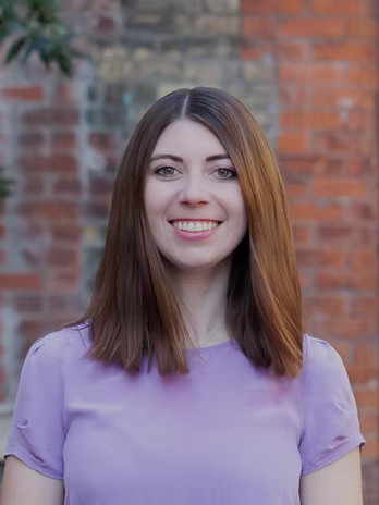 Julia smiling in a head-and-shoulders portrait in front of an old brick wall, framed by foliage.