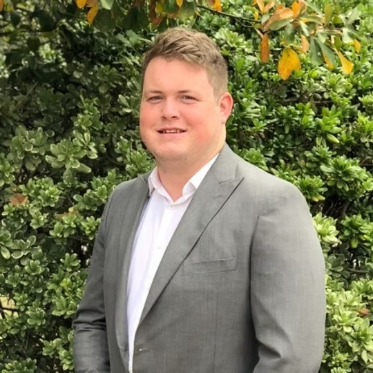 A man in a gray suit standing outdoors, smiling, with trees and foliage in the background.
