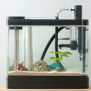 A wide, panoramic shot of a modern, high-tech freshwater aquarium setup that fills the majority of the frame. The tank sits on a wooden surface against a clean white background and features clear glass, sand substrate, porous rocks, live plants, and sophisticated filtration equipment including stainless steel pipes and tubing.