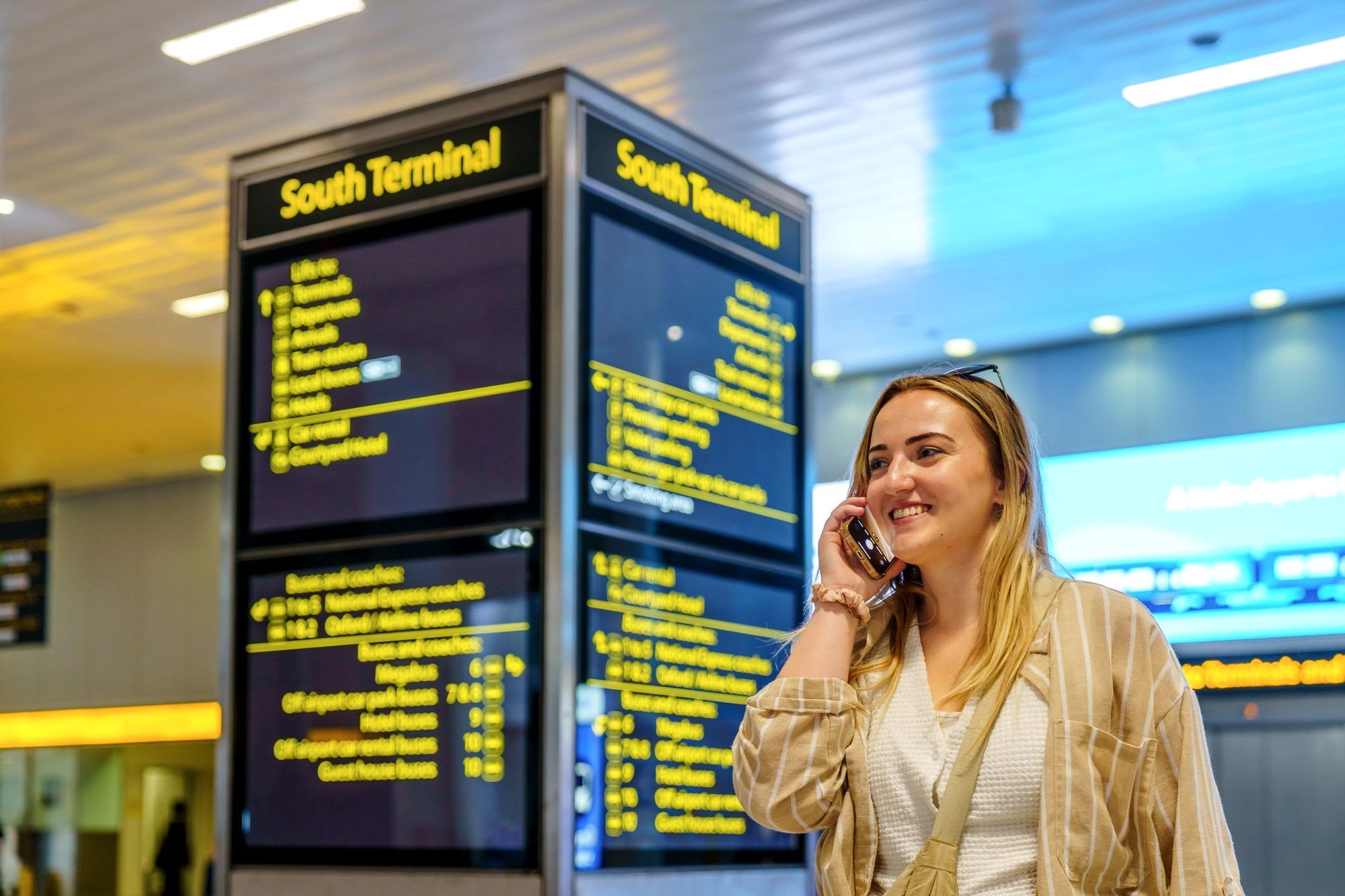 Person using a phone at an airport gate with a departures board behind them