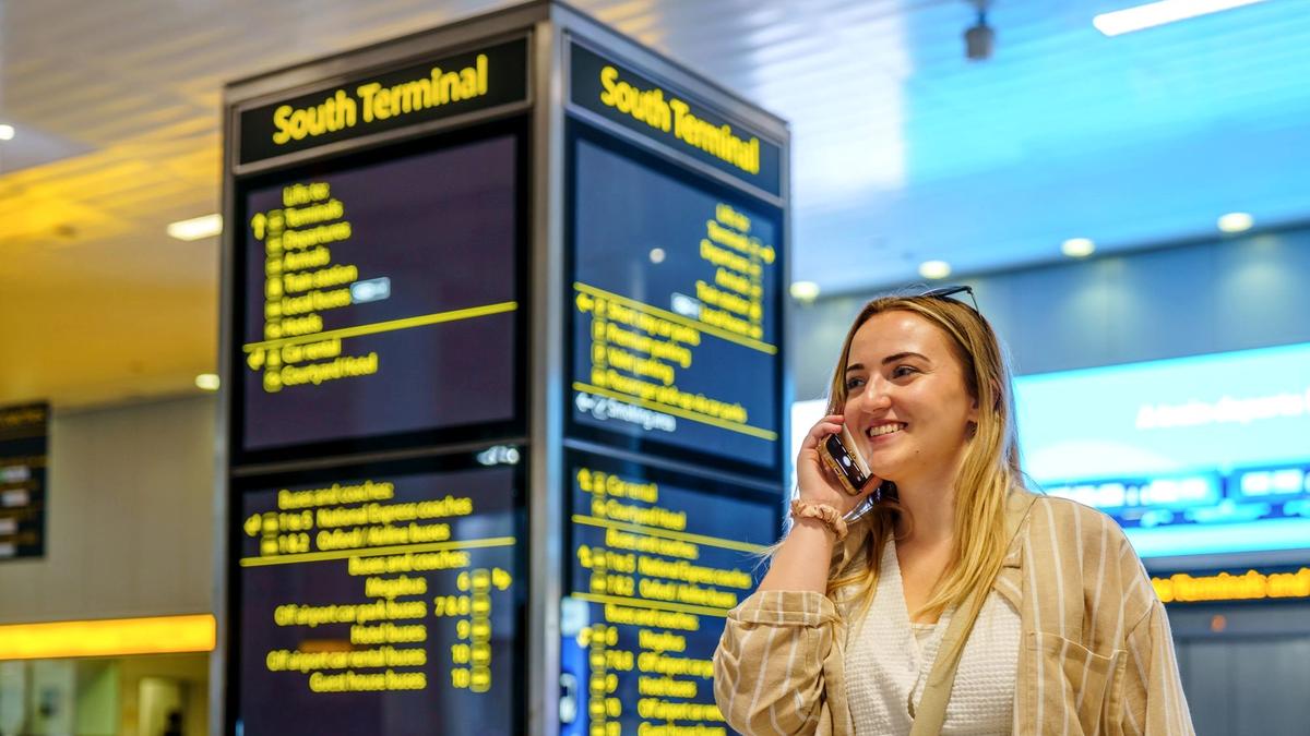 Person using a phone at an airport gate with a departures board behind them