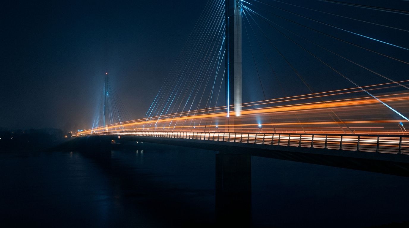 Cable-stayed bridge at night with orange light trails streaking across its span, fog softening the towers