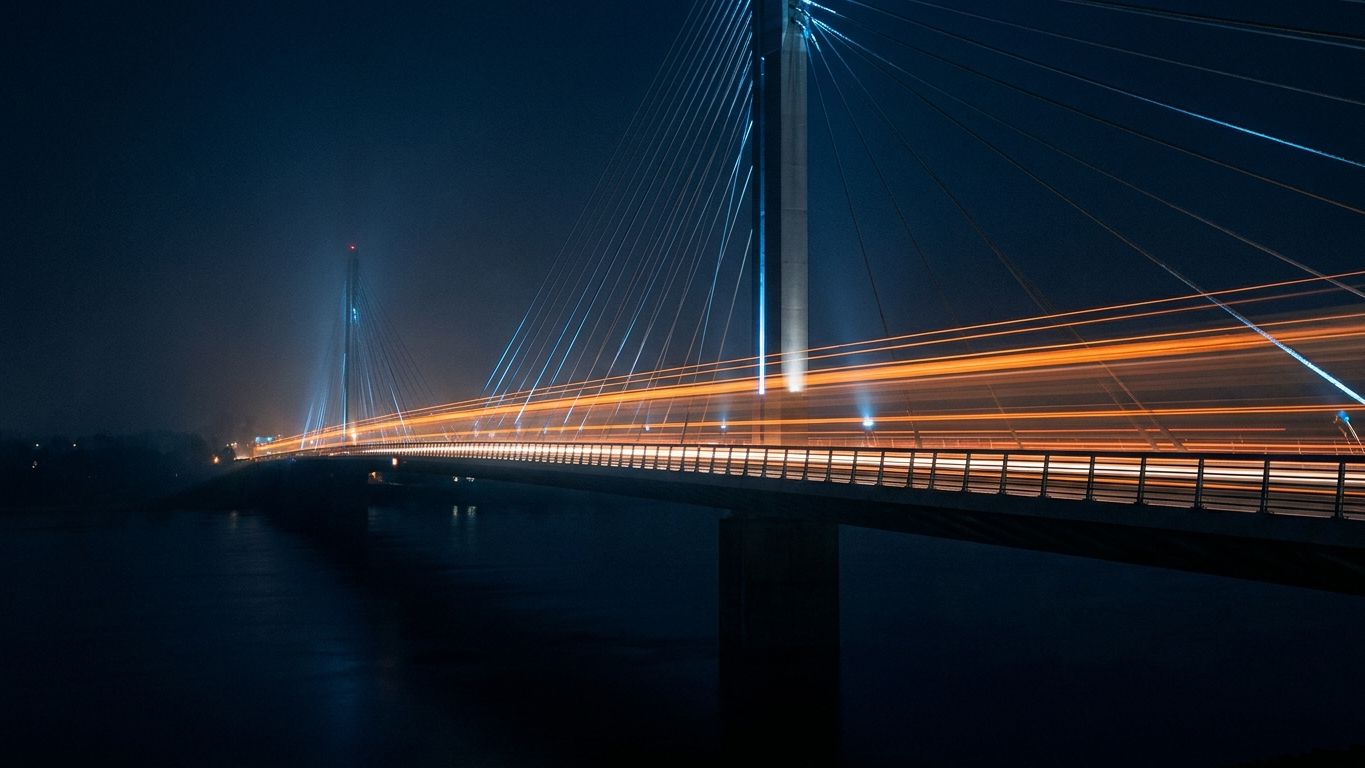 Cable-stayed bridge at night with orange light trails streaking across its span, fog softening the towers