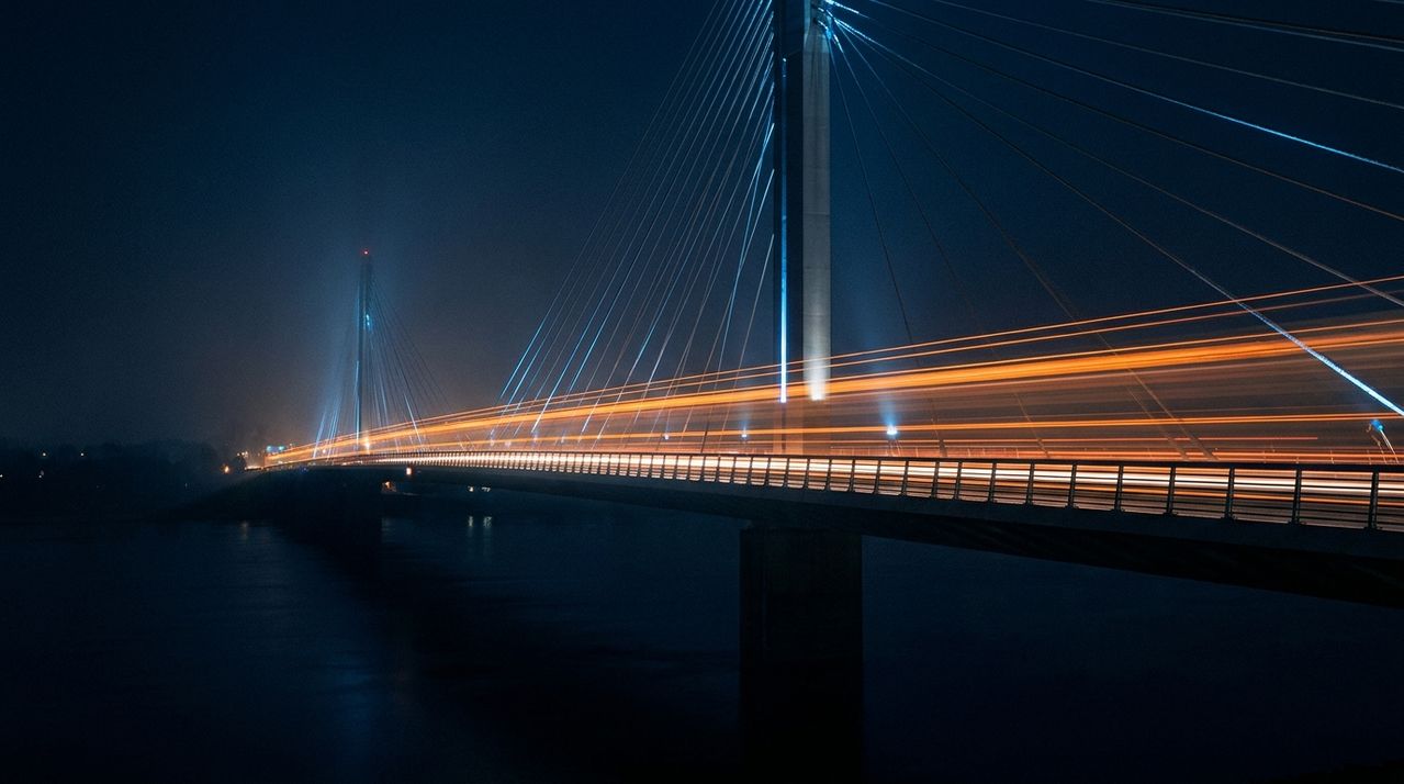 Cable-stayed bridge at night with orange light trails streaking across its span, fog softening the towers