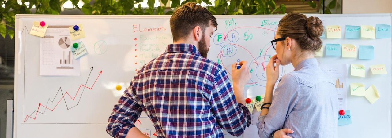 Two colleagues collaborating at a whiteboard covered in charts and sticky notes