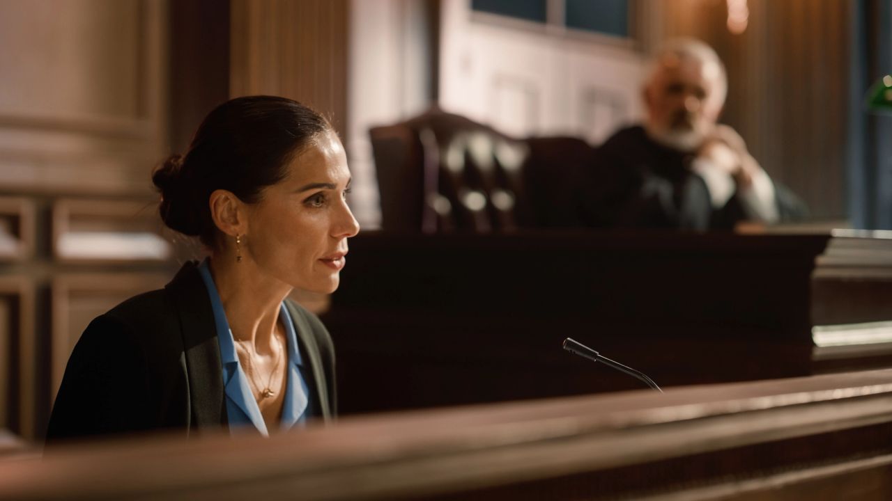 Woman speaking at a courtroom witness stand with a judge in the background