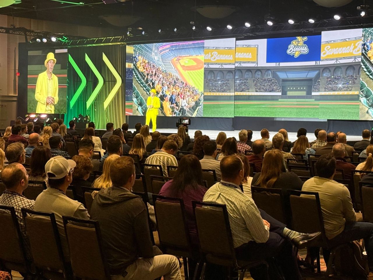 Conference keynote speaker in yellow suit presenting on stage to a seated audience