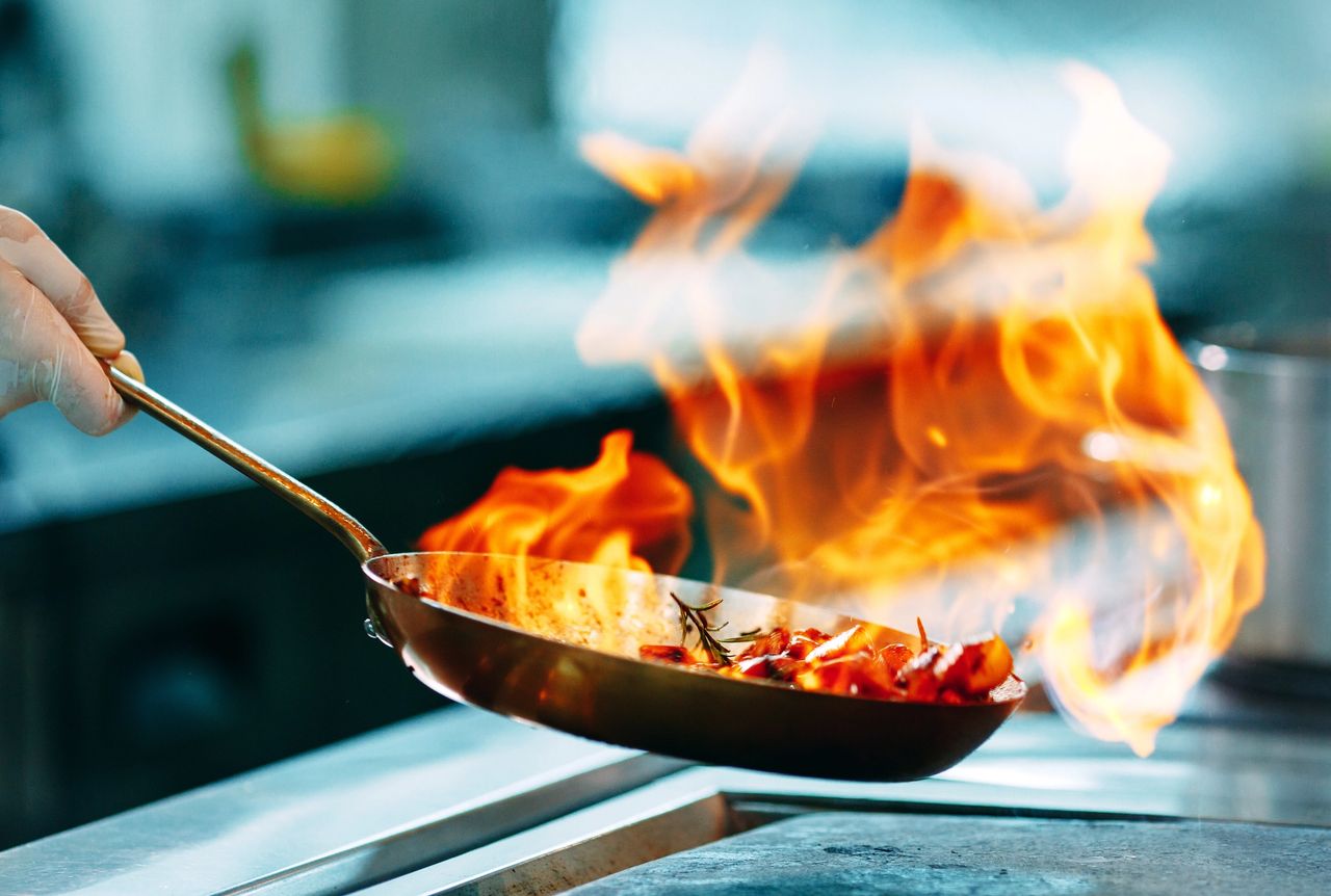 Chef flambing vegetables in a skillet with dramatic flames rising from the pan