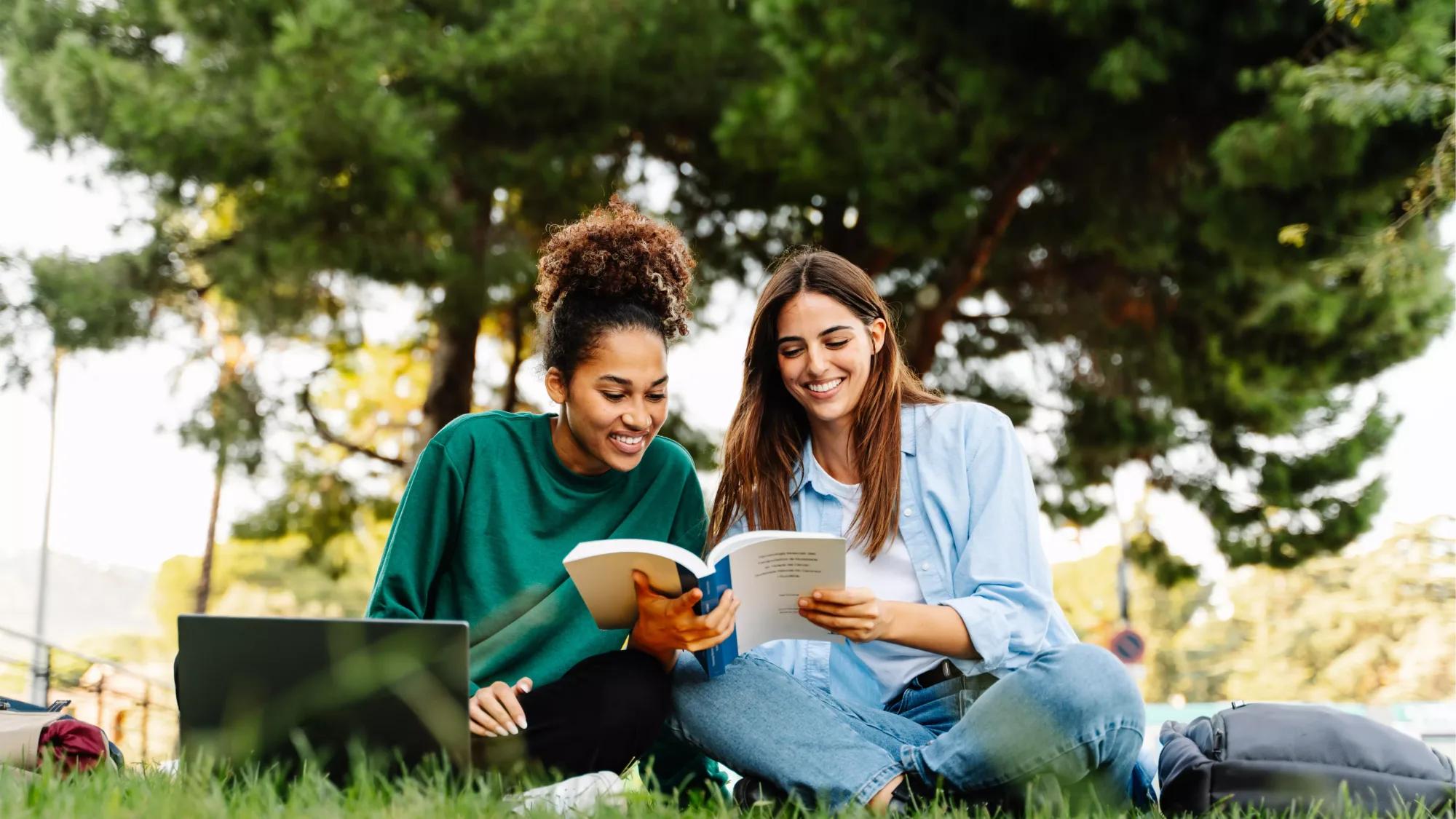 Two college students sitting on a campus lawn with a laptop and books, studying together under green trees