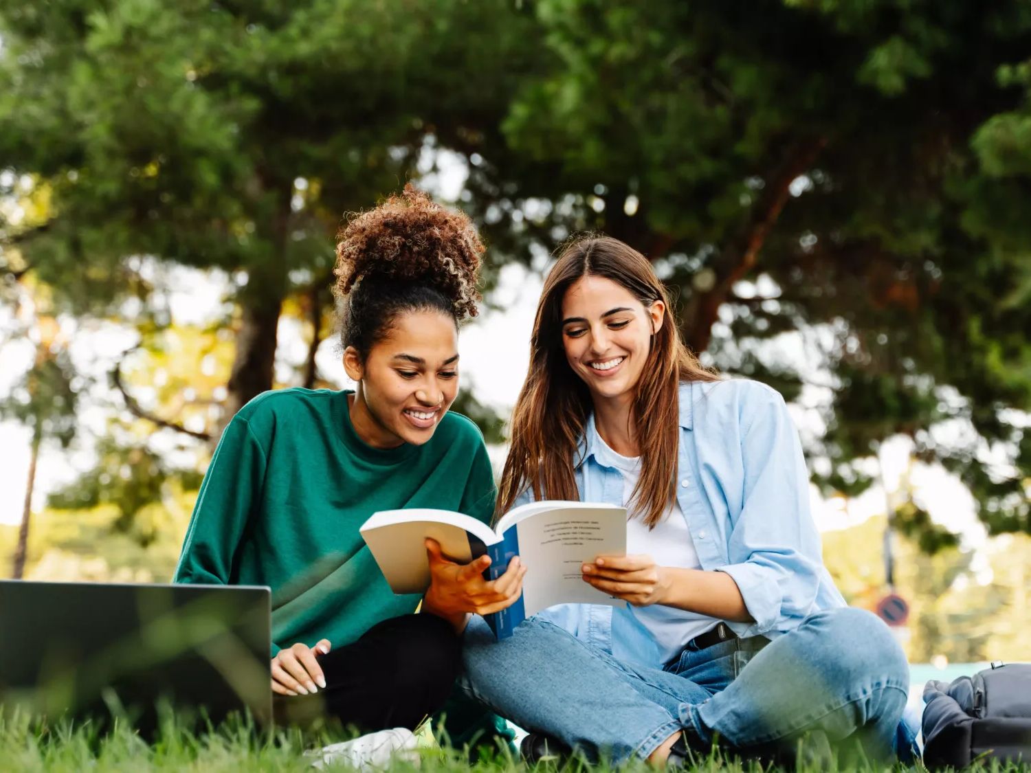 Two college students sitting on a campus lawn with a laptop and books, studying together under green trees