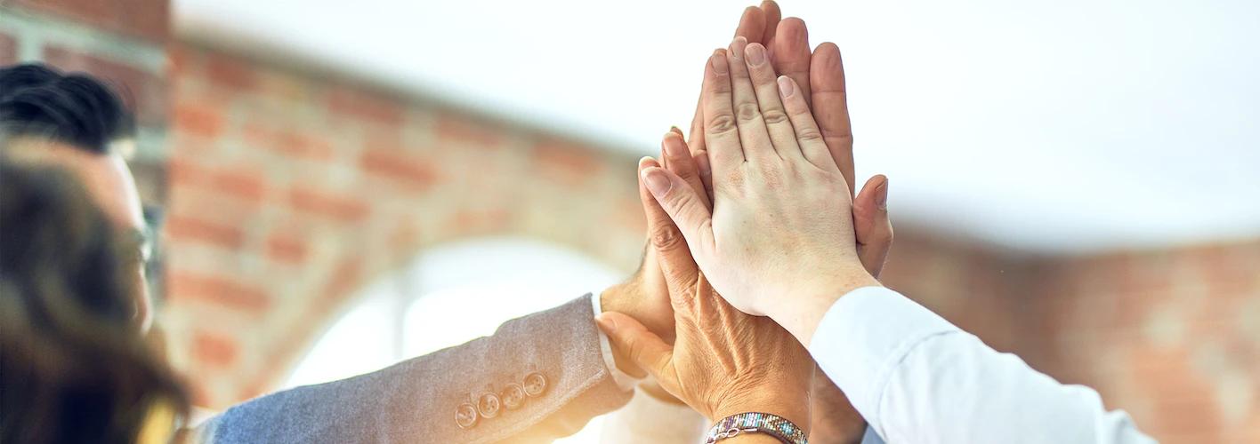 Team members joining hands together in a celebratory high-five against a brick wall backdrop