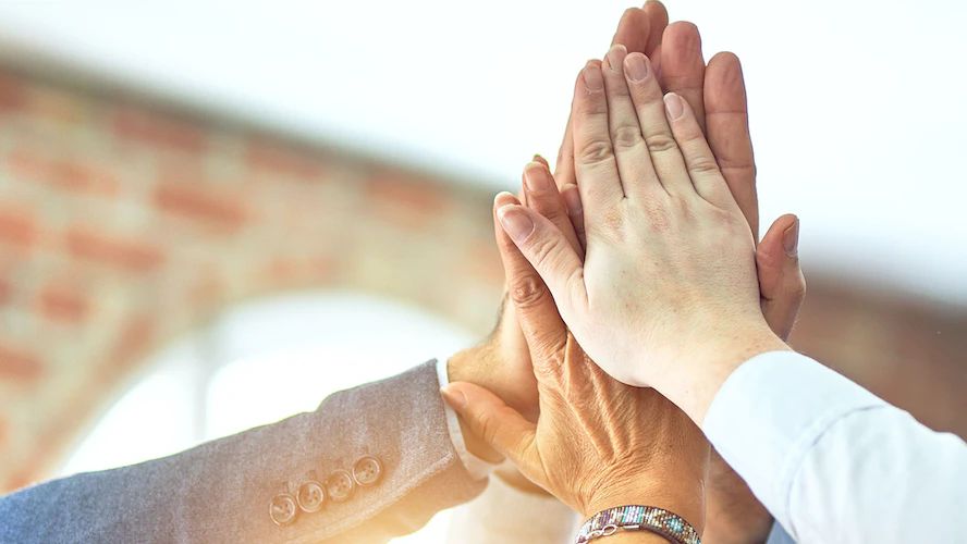 Team members joining hands together in a celebratory high-five against a brick wall backdrop