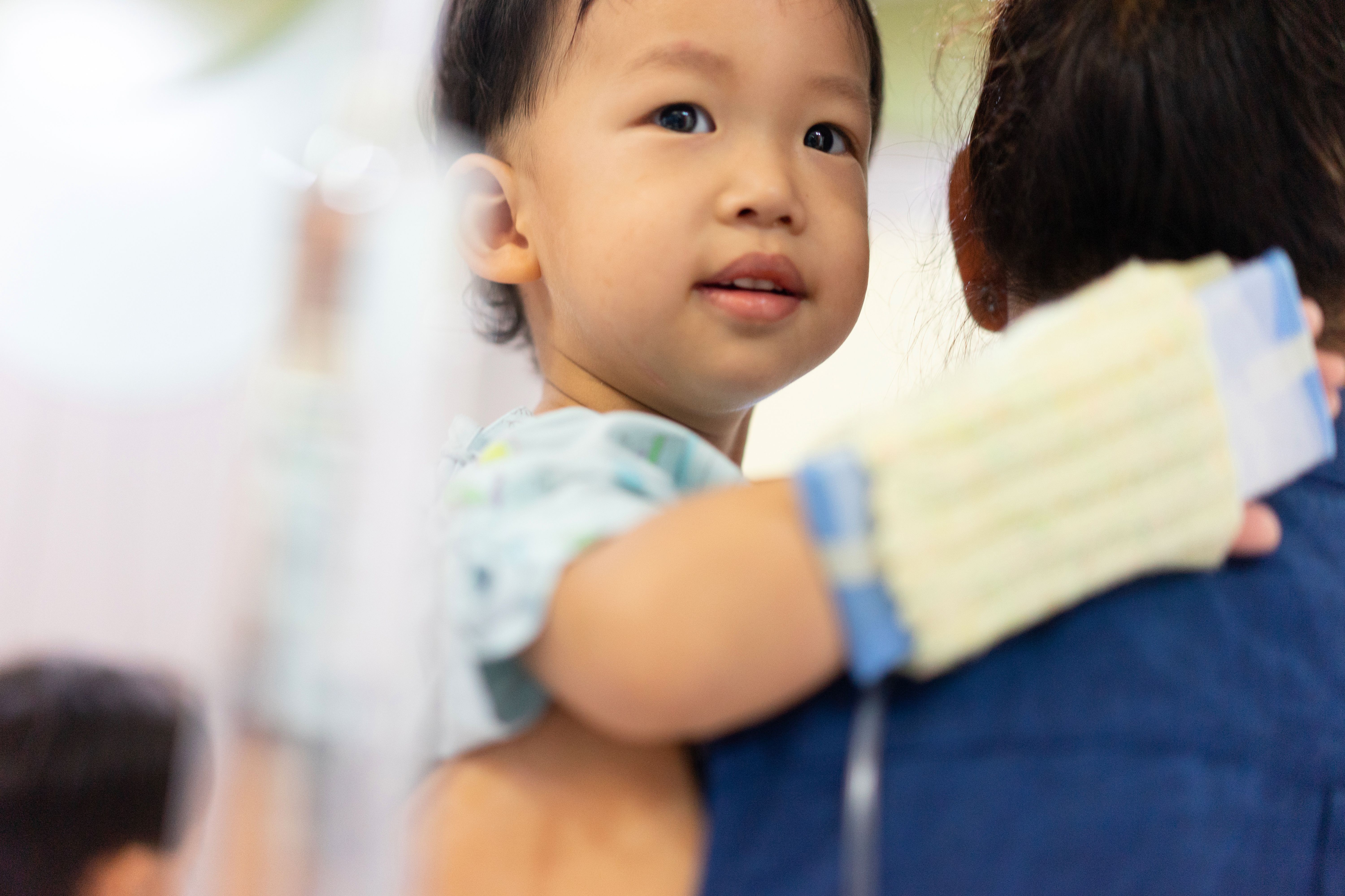 Young child with a hospital wristband held by a caregiver, looking over their shoulder with a gentle expression