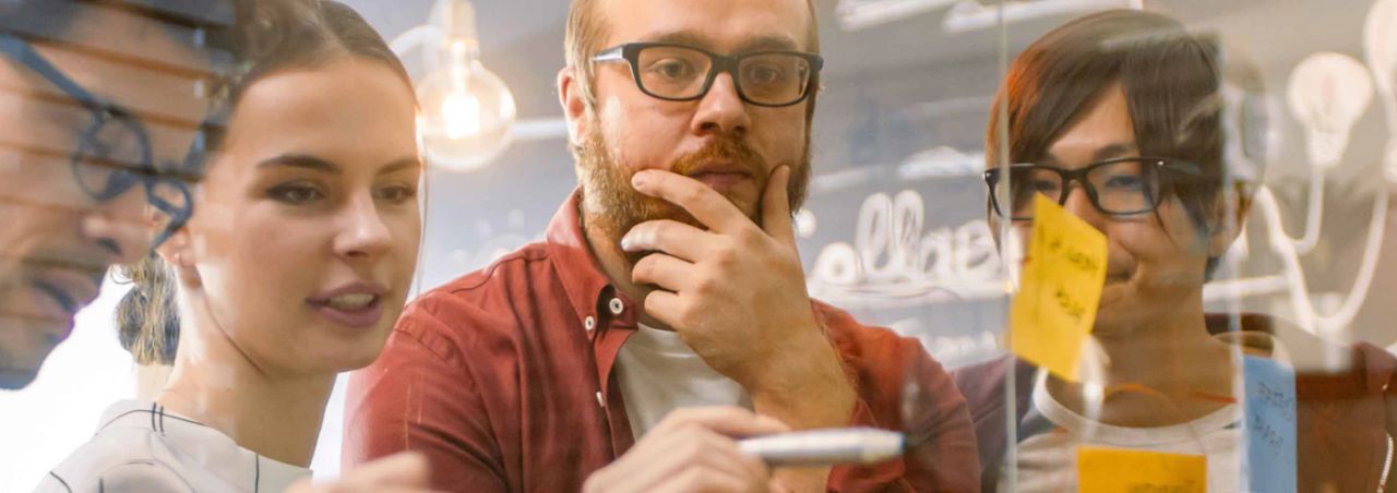 Three colleagues studying sticky notes and ideas written on a glass wall