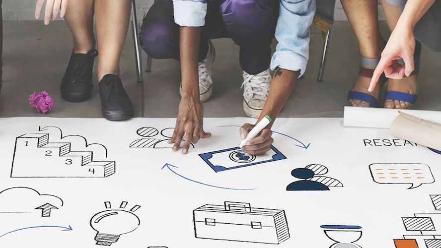 Diverse team drawing business and technology icons together on a large paper