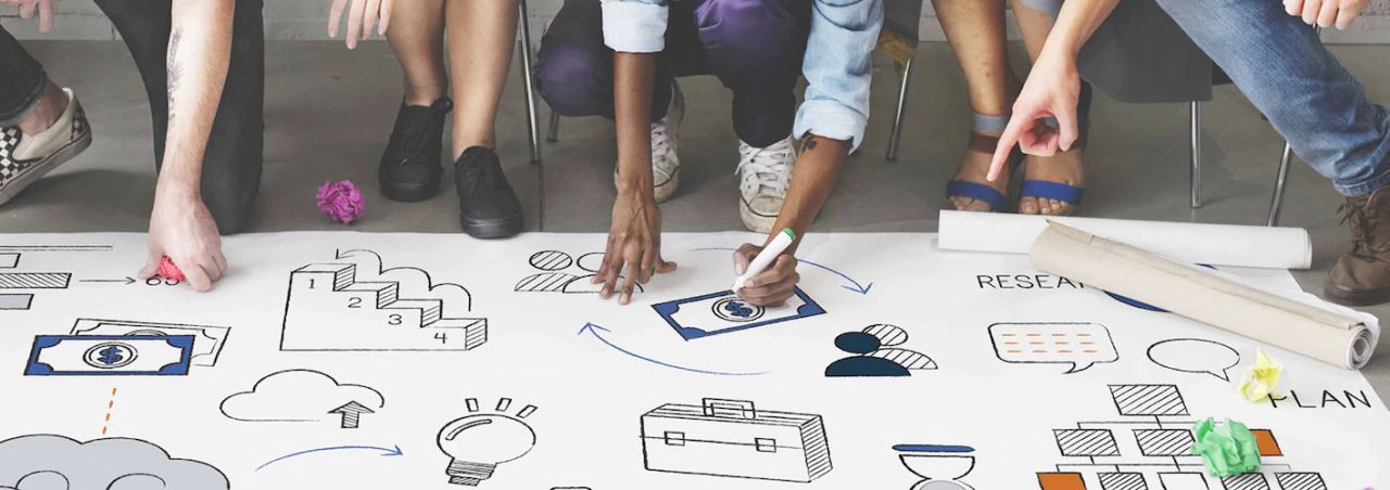 Diverse team drawing business and technology icons together on a large paper