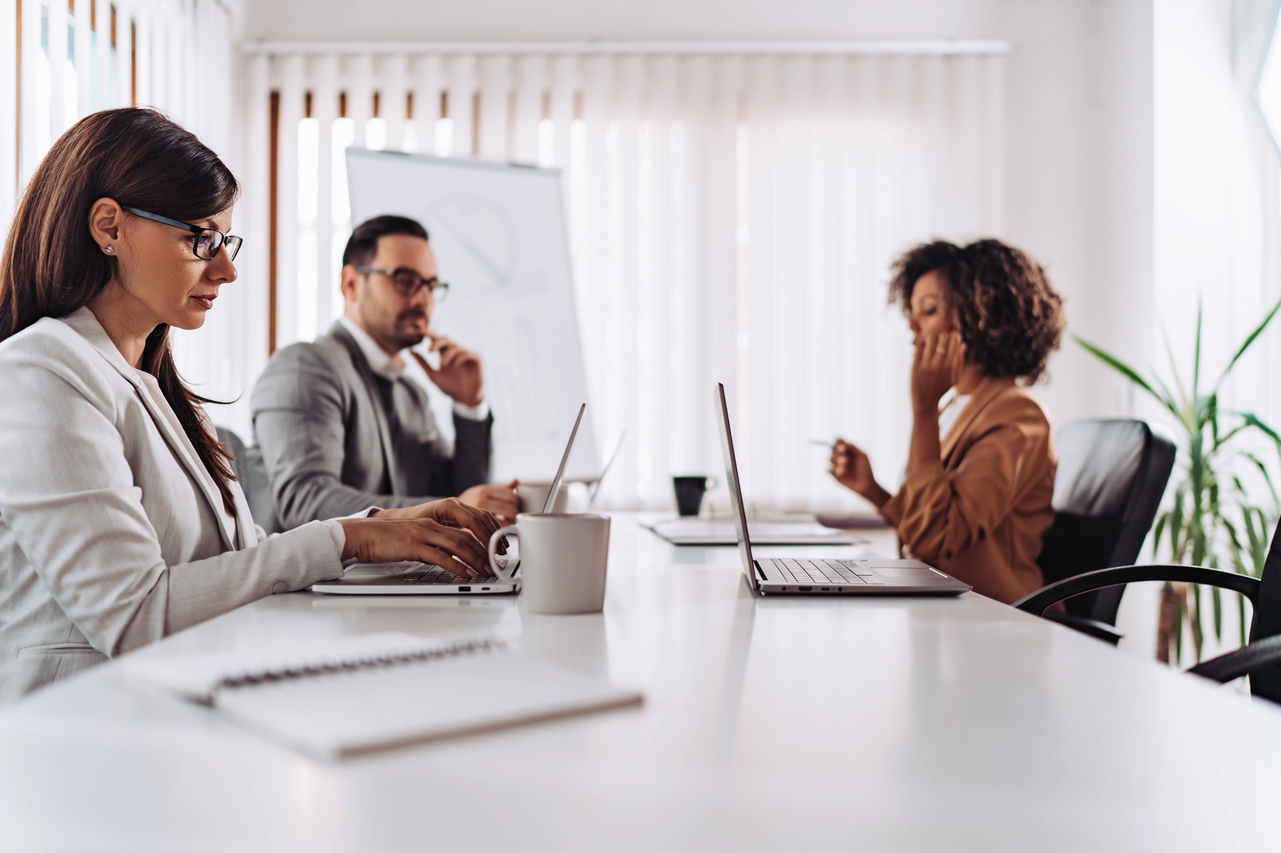 Three professionals working on laptops during a collaborative meeting in a modern office with a whiteboard