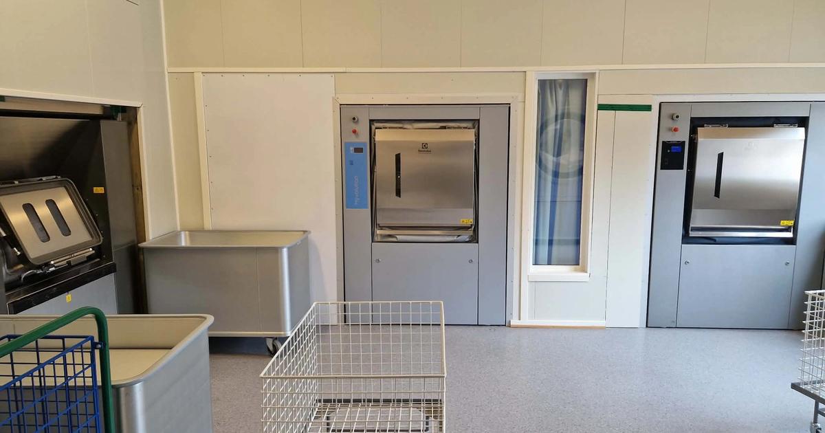 Interiør fra et vaskeri med en lang rekke hvite, industrielle vaskemaskiner eller tørketromler langs veggen. Rommet har et kjølig blåskjær, hvite flisgulv og lysrør i taket som skaper et symmetrisk perspektiv innover i lokalet.