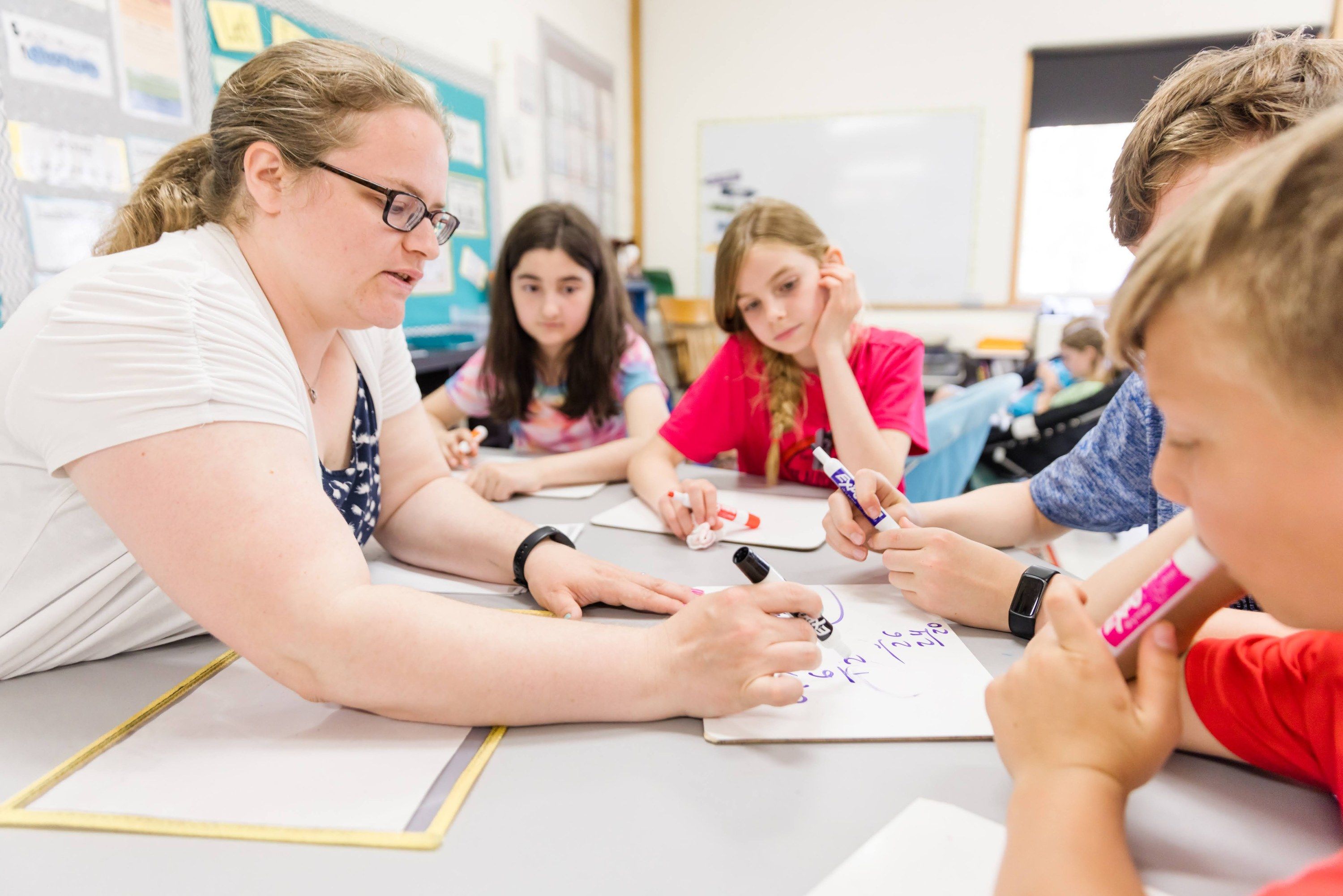A teacher works through math problems on whiteboards with a small group of students at a table.