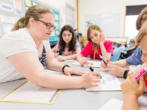 A teacher works through math problems on whiteboards with a small group of students at a table.